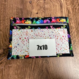 May include: A black zippered pouch with a colorful animal print border and a clear front panel decorated with red, blue, and green paint splatters. A white rectangle with the text "7x10" is on the front.