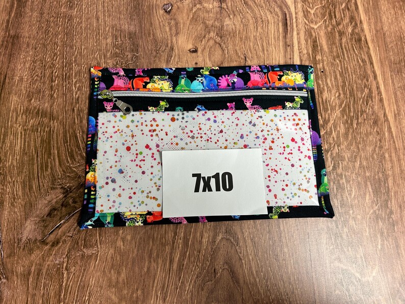 May include: A black zippered pouch with a colorful animal print border and a clear front panel decorated with red, blue, and green paint splatters. A white rectangle with the text "7x10" is on the front.