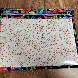 May include: A rectangular zippered pouch with a clear front and a black border. The pouch features a colorful dinosaur pattern along the top and bottom edges. The clear front is covered in a pattern of colorful paint splatters.