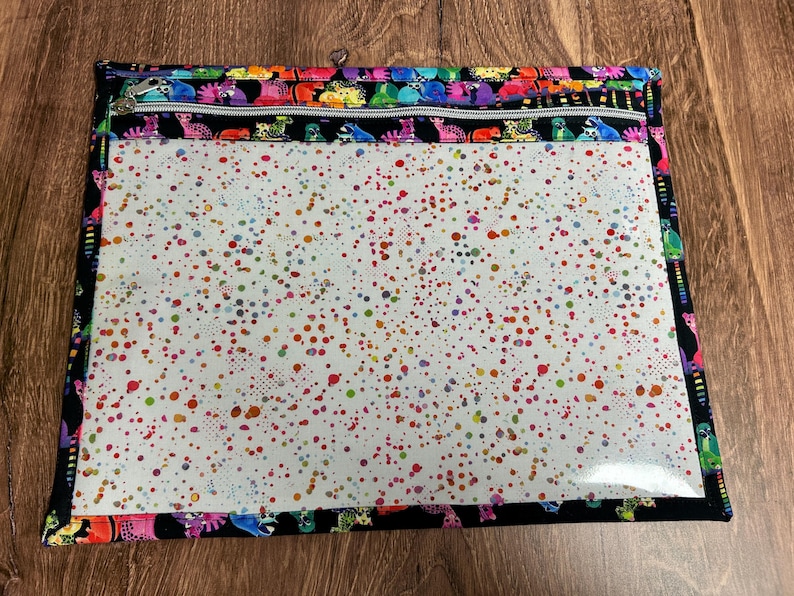 May include: A rectangular zippered pouch with a clear front and a black border. The pouch features a colorful dinosaur pattern along the top and bottom edges. The clear front is covered in a pattern of colorful paint splatters.