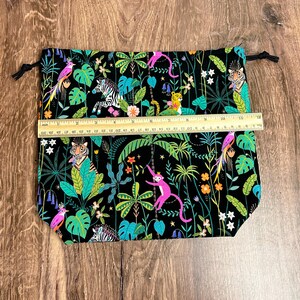 May include: A black drawstring bag with a jungle-themed print. The fabric features colorful illustrations of tigers, zebras, monkeys, and parrots amidst lush green foliage. The bag measures approximately 30 cm wide.
