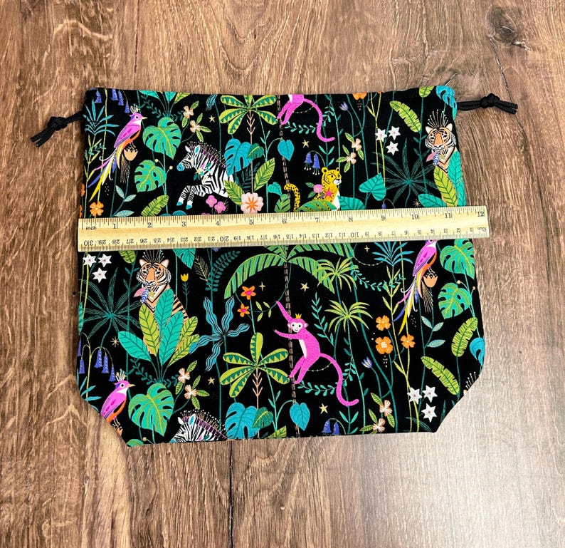 May include: A black drawstring bag with a jungle-themed print. The fabric features colorful illustrations of tigers, zebras, monkeys, and parrots amidst lush green foliage. The bag measures approximately 30 cm wide.
