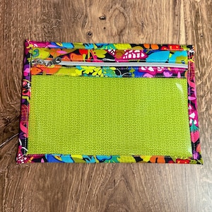 Puede incluir: Una bolsa rectangular con cremallera y ventana transparente. La bolsa presenta un vibrante estampado floral en rosa, amarillo, azul y morado. La ventana es de color verde brillante con un patr&oacute;n repetitivo. La cremallera es plateada.
