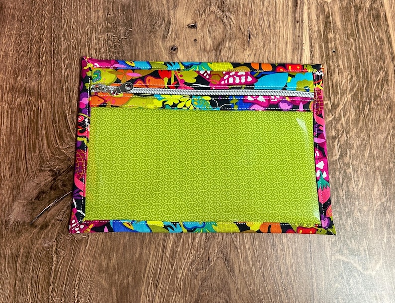 Puede incluir: Una bolsa rectangular con cremallera y ventana transparente. La bolsa presenta un vibrante estampado floral en rosa, amarillo, azul y morado. La ventana es de color verde brillante con un patr&oacute;n repetitivo. La cremallera es plateada.