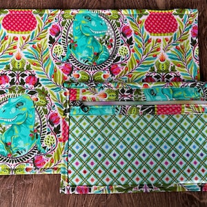 May include: A green and white fabric placemat with a dinosaur pattern. A smaller green and white fabric pouch with a zipper is attached to the placemat.