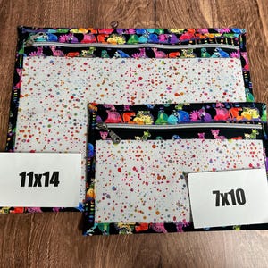 May include: Two zippered pouches in different sizes. The larger pouch is 11x14 inches, and the smaller is 7x10 inches. Both have a black border with a colorful creature pattern and a clear front with a speckled design. The zippers are silver.