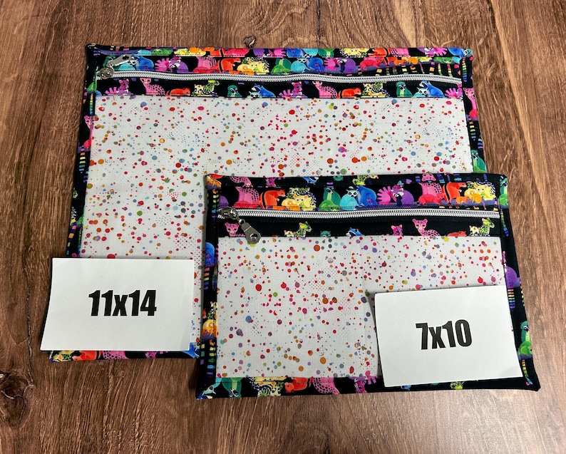 May include: Two zippered pouches in different sizes. The larger pouch is 11x14 inches, and the smaller is 7x10 inches. Both have a black border with a colorful creature pattern and a clear front with a speckled design. The zippers are silver.