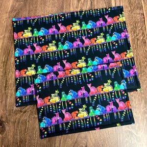 May include: Two black placemats with a repeating pattern of colorful, stylized animals. The animals are in shades of pink, purple, blue, green, yellow, and orange. The background is black, and the design has a rainbow stripe effect.