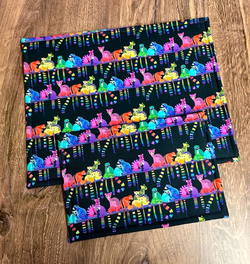 May include: Two black placemats with a repeating pattern of colorful, stylized animals. The animals are in shades of pink, purple, blue, green, yellow, and orange. The background is black, and the design has a rainbow stripe effect.