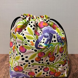 May include: A drawstring bag with a colorful pattern of purple monkeys, cherries, and green leaves on a cream background. The bag has black drawstring cords and is sitting on a wooden surface.