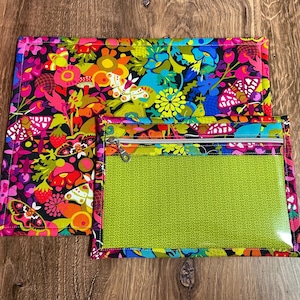 Puede incluir: Un conjunto con estampado floral colorido que incluye una alfombrilla rectangular grande y una bolsa m&aacute;s peque&ntilde;a con cremallera. La alfombrilla y la bolsa tienen un fondo negro con flores vibrantes, mariposas y fresas en tonos de rosa, naranja, amarillo y azul. La bolsa tiene un frente transparente y un interior verde estampado.