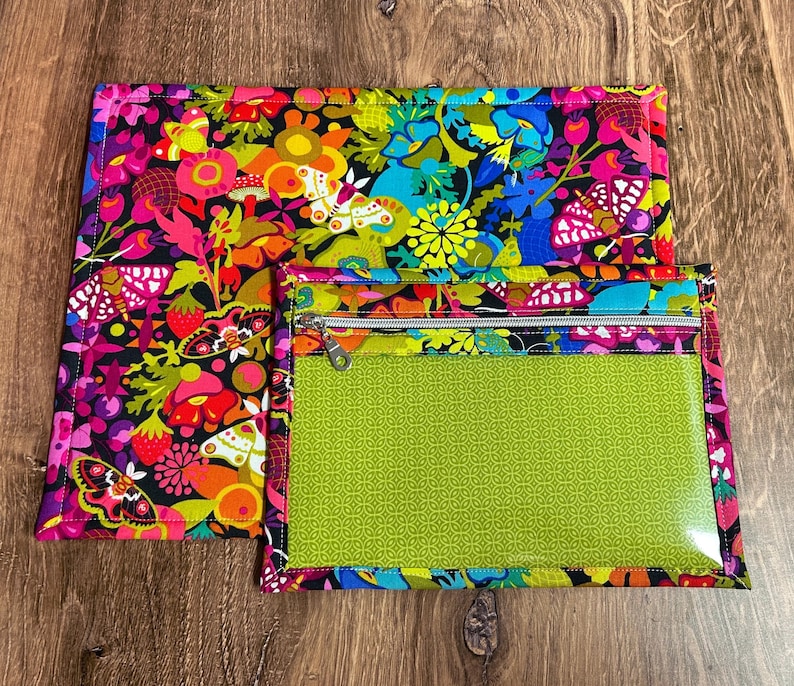 Puede incluir: Un conjunto con estampado floral colorido que incluye una alfombrilla rectangular grande y una bolsa m&aacute;s peque&ntilde;a con cremallera. La alfombrilla y la bolsa tienen un fondo negro con flores vibrantes, mariposas y fresas en tonos de rosa, naranja, amarillo y azul. La bolsa tiene un frente transparente y un interior verde estampado.