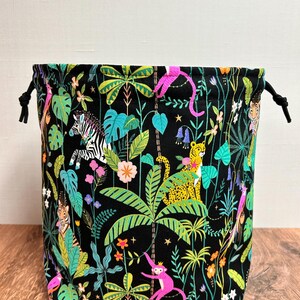 May include: A black drawstring bag with a jungle-themed pattern. The fabric is adorned with colourful illustrations of animals, including zebras, monkeys, and leopards, set against a backdrop of green foliage and flowers. It has black drawstrings.