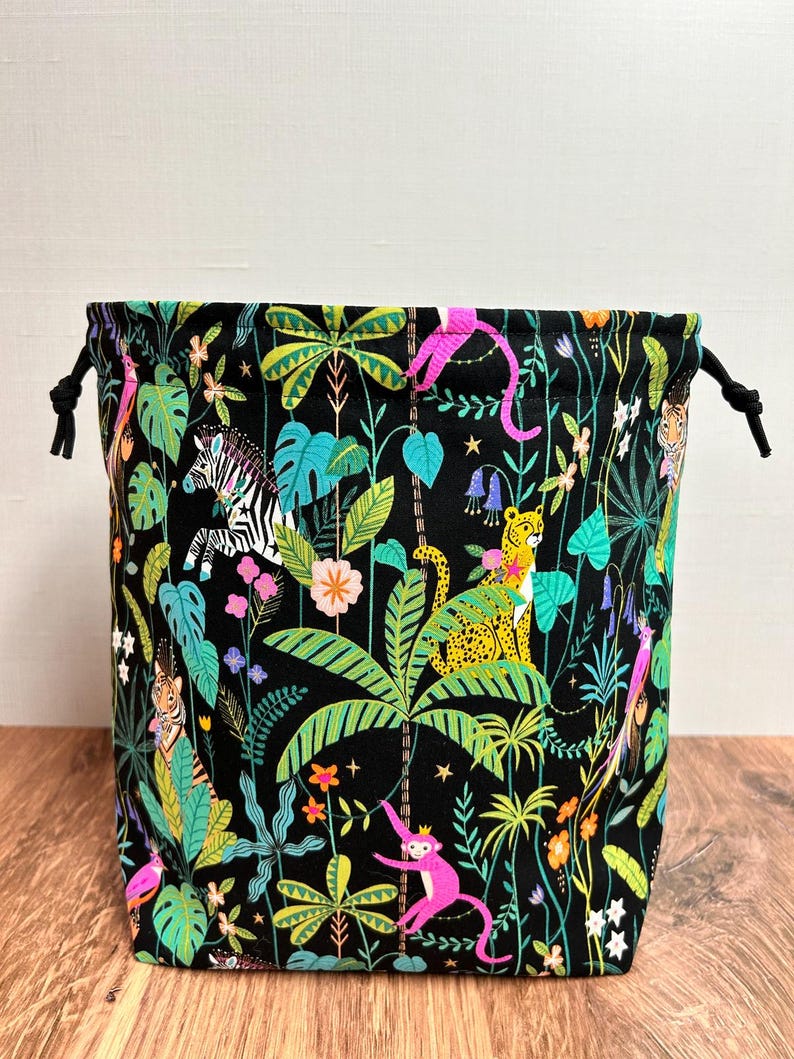 May include: A black drawstring bag with a jungle-themed pattern. The fabric is adorned with colourful illustrations of animals, including zebras, monkeys, and leopards, set against a backdrop of green foliage and flowers. It has black drawstrings.