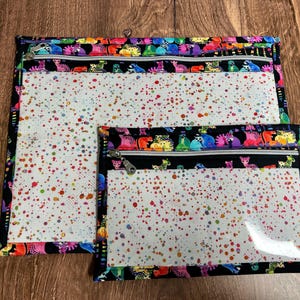 May include: Two zippered pouches with a colorful dinosaur pattern border. The pouches have clear plastic fronts with a white background speckled with red, yellow, green, and blue dots. The zippers are silver.