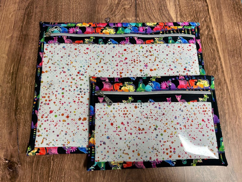 May include: Two zippered pouches with a colorful dinosaur pattern border. The pouches have clear plastic fronts with a white background speckled with red, yellow, green, and blue dots. The zippers are silver.