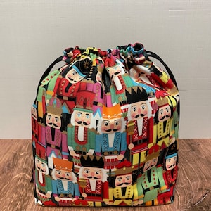 May include: A drawstring bag made of fabric with a colorful pattern of nutcracker figurines. The bag has black drawstrings and a black bottom.