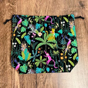 May include: Black drawstring bag with a vibrant jungle print. The design features colourful parrots, monkeys, zebras, and tigers amidst lush green foliage and flowers. The bag has a black drawstring closure.