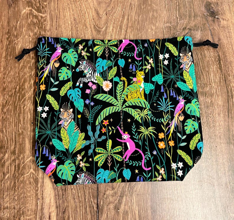 May include: Black drawstring bag with a vibrant jungle print. The design features colourful parrots, monkeys, zebras, and tigers amidst lush green foliage and flowers. The bag has a black drawstring closure.