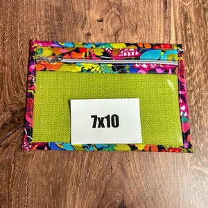 Puede incluir: Una bolsa rectangular con una parte delantera transparente y un borde con estampado floral colorido. La bolsa tiene un cierre de cremallera y una tarjeta blanca con el texto "7x10". La bolsa mide aproximadamente 18 cm por 25 cm.