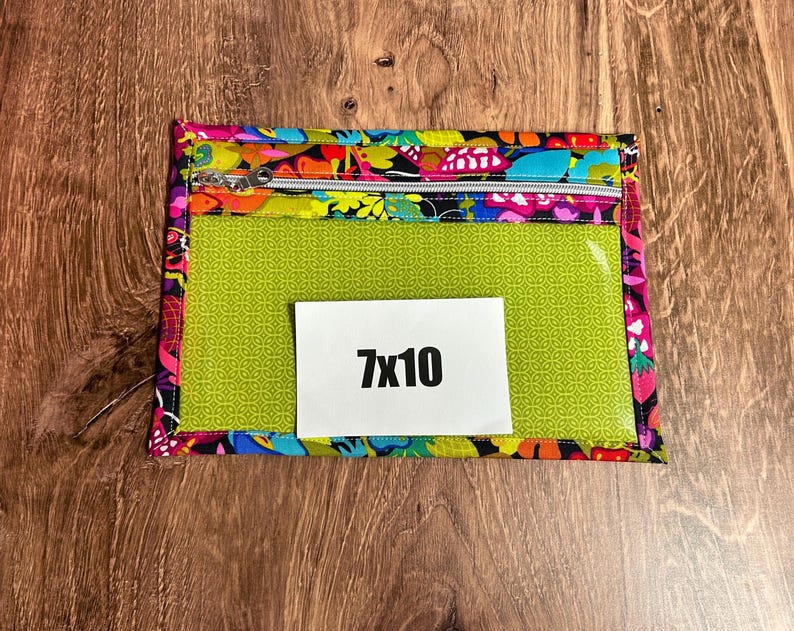 Puede incluir: Una bolsa rectangular con una parte delantera transparente y un borde con estampado floral colorido. La bolsa tiene un cierre de cremallera y una tarjeta blanca con el texto "7x10". La bolsa mide aproximadamente 18 cm por 25 cm.