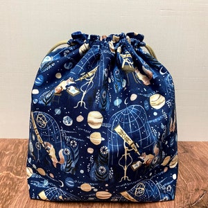 May include: A navy blue drawstring bag with a celestial pattern featuring planets, telescopes, and constellations. The bag is made of fabric and has a beige drawstring closure. The bag is sitting on a wooden surface.
