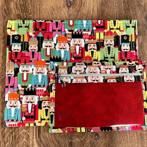 May include: A red zippered pouch with a clear plastic window. The pouch is decorated with a colorful pattern of nutcracker figurines.