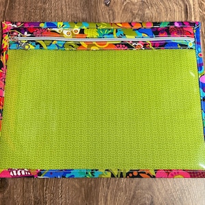 Puede incluir: Bolsa rectangular con frente transparente y borde con estampado floral colorido. El interior es verde brillante con un patr&oacute;n repetitivo. La cremallera plateada y el borde presentan dise&ntilde;os florales rosas, azules, naranjas y negros.