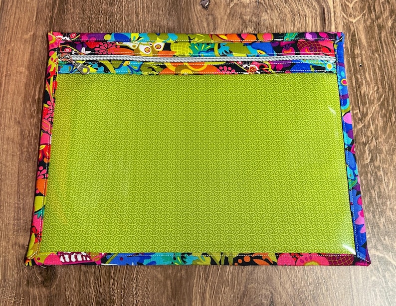 Puede incluir: Bolsa rectangular con frente transparente y borde con estampado floral colorido. El interior es verde brillante con un patr&oacute;n repetitivo. La cremallera plateada y el borde presentan dise&ntilde;os florales rosas, azules, naranjas y negros.