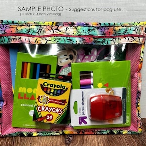 Puede incluir: Una bolsa de vinilo transparente, de aproximadamente 28 cm x 36 cm, con un borde de tela estampada. La bolsa contiene &uacute;tiles escolares, incluyendo crayones, l&aacute;pices de colores y un sacapuntas. El texto "SAMPLE PHOTO - Suggestions for bag use" es visible.