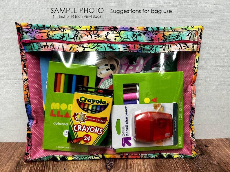 Puede incluir: Una bolsa de vinilo transparente, de aproximadamente 28 cm x 36 cm, con un borde de tela estampada. La bolsa contiene &uacute;tiles escolares, incluyendo crayones, l&aacute;pices de colores y un sacapuntas. El texto "SAMPLE PHOTO - Suggestions for bag use" es visible.