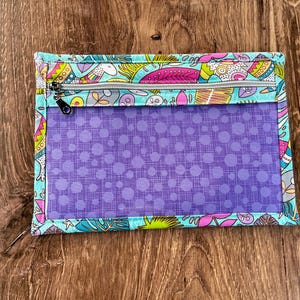 Puede incluir: Una bolsa rectangular con cremallera, panel frontal transparente y borde estampado colorido. El borde presenta un fondo turquesa con dise&ntilde;os florales y de hojas rosas, amarillas y moradas. El interior es morado con un estampado de lunares.