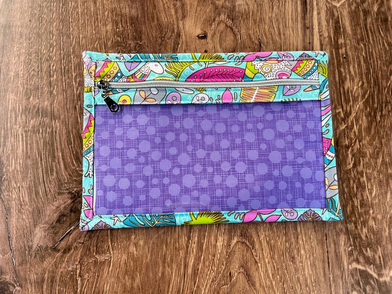 Puede incluir: Una bolsa rectangular con cremallera, panel frontal transparente y borde estampado colorido. El borde presenta un fondo turquesa con dise&ntilde;os florales y de hojas rosas, amarillas y moradas. El interior es morado con un estampado de lunares.