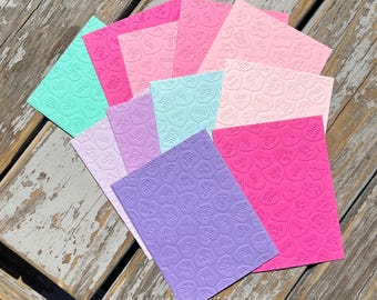 May include: A collection of rectangular cards in various pastel and vibrant colors, including pink, purple, blue, and green, each featuring an embossed pattern of small heart-shaped candies with phrases like LUV, XOXO, LOL, and CALL ME.
