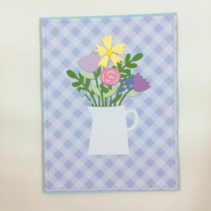 May include: A white card with a blue and white checked pattern. A white jug with a handle is filled with a bouquet of paper flowers in shades of pink, purple, yellow, and blue.