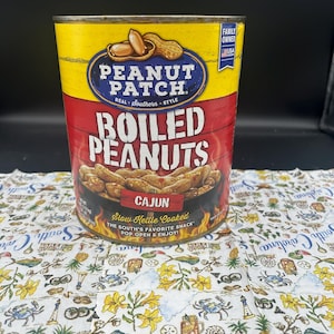 May include: A can of Peanut Patch boiled peanuts with a yellow top and red bottom. The label reads "Boiled Peanuts Cajun" with the text "Slow Kettle Cooked" and "Pop Open & Enjoy!". The can is on a patterned cloth.