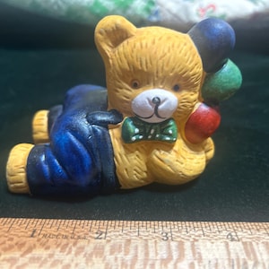 May include: A ceramic teddy bear figurine, lying down, wearing blue overalls and a green bow tie. The bear holds three balloons in red, green, and blue. The bear is yellow with a white snout.
