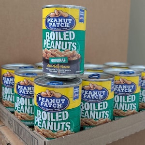 May include: A stack of canned boiled peanuts, with the brand name "Peanut Patch" prominently displayed. The cans are yellow, green, and blue, with text that reads "Original" and "The South's Favorite Snack". The cans are in a cardboard tray.