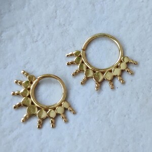 Puede incluir: Dos anillos de tabique dorados, cada uno con un diseño de sol y pequeños detalles circulares. Se presentan sobre una superficie azul claro texturizada. Los anillos dorados contrastan con el fondo azul claro.