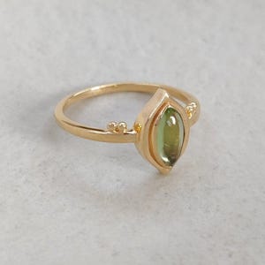 May include: A gold ring with a teardrop-shaped, olive-green gemstone. The ring has a delicate band and small decorative details on either side of the gemstone. The ring is set against a white background.