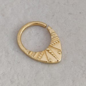 Puede incluir: Un anillo de tabique dorado con un diseño abierto. El anillo presenta un elemento decorativo de forma triangular con patrones geométricos grabados y pequeños detalles de puntos. La joya está sobre un fondo neutro.