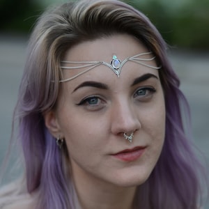 May include: Silver-toned headpiece with a central opal-like stone and delicate chain accents. The image also shows a matching necklace and nose jewelry. The person has purple hair and is wearing a black off-the-shoulder top.