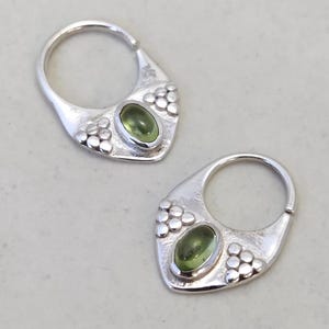 Puede incluir: Un par de pendientes de aro de plata con una piedra preciosa verde engastada en cada uno. Los pendientes tienen un diseño texturizado alrededor de la piedra preciosa.