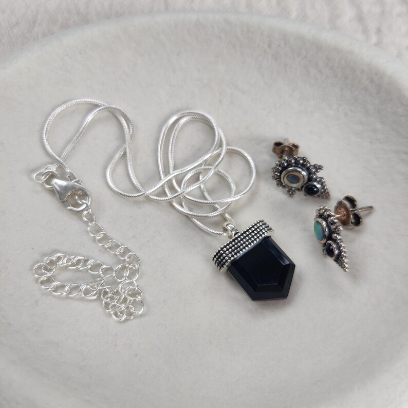 Silver earrings and silver necklace with black onyx crystal pendant, with a snake chain, displayed on a jewelry plate.
