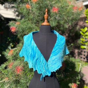 May include: A hand-knitted triangular shawl in variegated shades of bright turquoise and sky blue. The accessory features a textured ribbed stitch pattern with vertical eyelet details along the edges, creating a soft, draped, and wavy appearance.