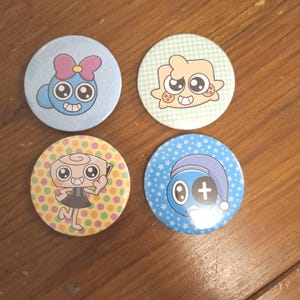 Pode incluir: Quatro botões redondos com desenhos de personagens de desenhos animados. Os botões apresentam fundos azul claro, amarelo e verde com bolinhas, estrelas e padrões xadrez. Cada botão tem uma ilustração de personagem única.