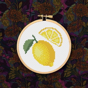 May include: A cross-stitch embroidery hoop showing a yellow lemon and a slice with a green leaf. The design is on white fabric, set against a floral patterned background with dark purple, orange, and green hues.