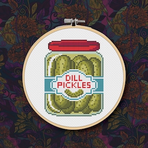 Puede incluir: Un aro de bordado de punto de cruz con un diseño de un frasco de vidrio lleno de pepinillos encurtidos. El frasco tiene una tapa roja y una etiqueta azul que dice "Dill Pickles".