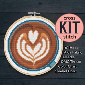 May include: A cross stitch kit with a coffee latte design, showing a heart-shaped foam pattern. The kit includes a 15 cm hoop, Aida fabric, a needle, DMC thread, a colour chart, and a symbol chart. The design uses brown, white, and blue threads.