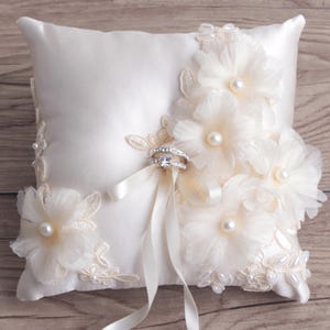 May include: A white satin ring pillow with delicate lace and pearl embellishments. Two silver rings are placed on the pillow.